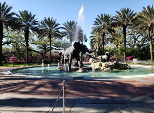 A fountain featuring an elephant statue surrounded by palm trees and colorful flowers in a sunny park setting.