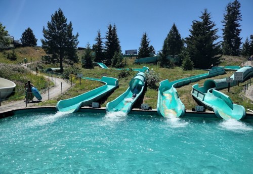 A water park scene featuring multiple turquoise water slides and a clear blue pool under a sunny sky.
