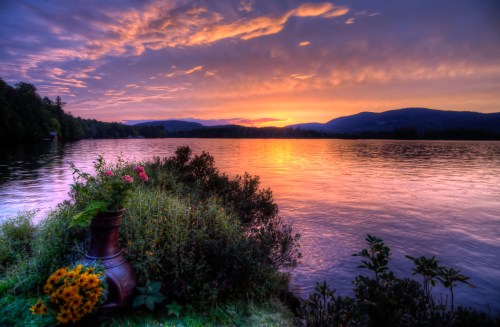 A serene lakeside scene at sunset, with vibrant colors reflecting on the water and flowers in the foreground.