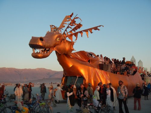 A large, golden dragon sculpture on a vehicle surrounded by people at a desert festival during sunset.