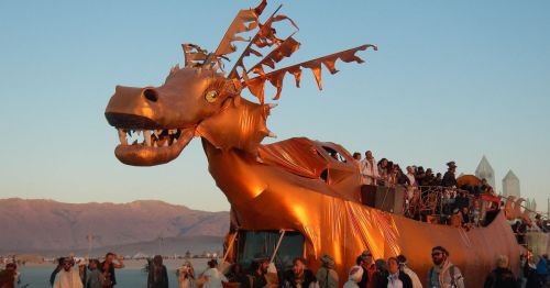 A large, golden dragon sculpture with people on top, set against a desert landscape at sunset.