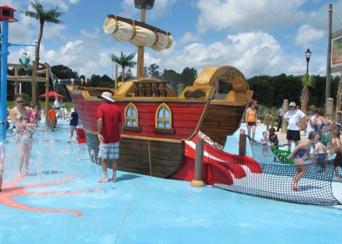 A colorful pirate ship play structure in a water park, with children playing and splashing around in the water.