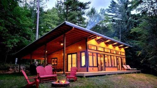 A modern cabin in the woods with a fire pit and red chairs, surrounded by trees and a twilight sky.