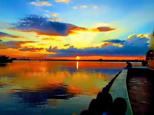 A vibrant sunset over a calm river, with colorful clouds reflecting on the water and a silhouette of a pier.