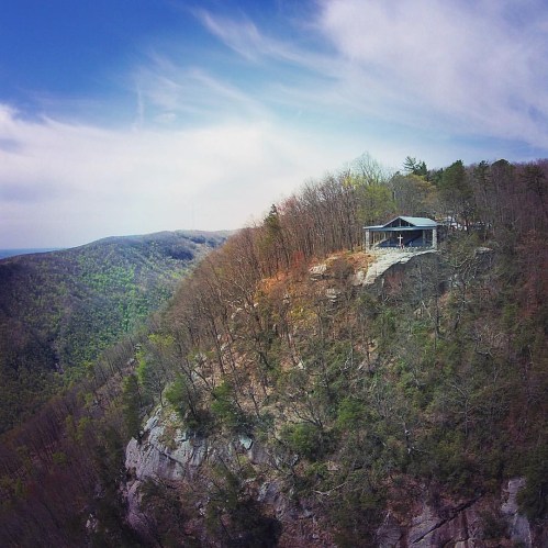A scenic view of a house perched on a rocky cliff, surrounded by lush green mountains and a clear blue sky.