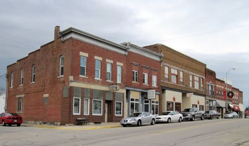 A small town street featuring brick buildings, shops, and parked cars under a cloudy sky.