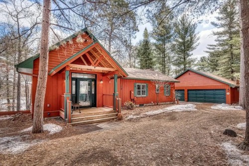 A charming red cabin surrounded by trees, featuring a front porch and an attached garage. Snow is visible on the ground.