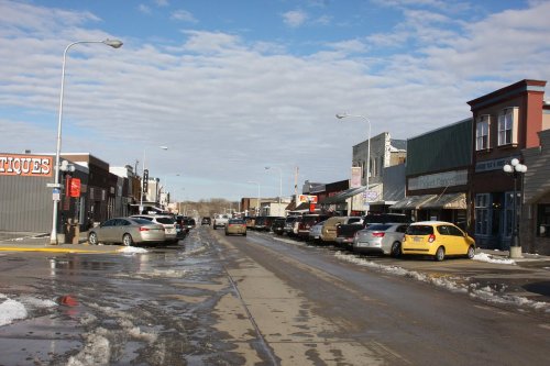 A street lined with shops and parked cars, featuring a mix of snow and puddles under a cloudy sky.