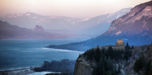 A scenic view of a river valley surrounded by mountains, with a historic building perched on a cliff.