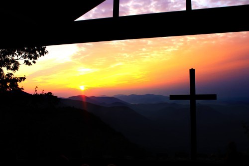 Silhouette of a cross against a vibrant sunset over mountains, viewed through a window.