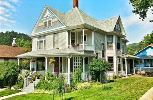 A charming two-story house with a decorative roof, surrounded by greenery and flowers in a sunny neighborhood.