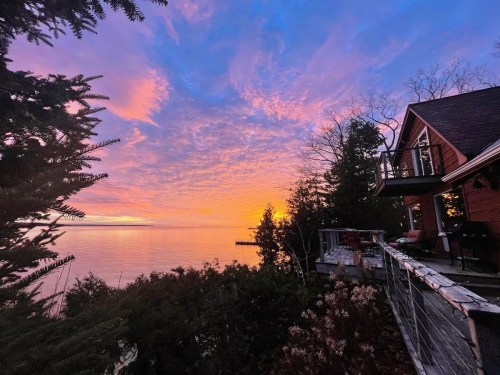 A vibrant sunset over a calm lake, with colorful clouds and a cozy cabin surrounded by trees.