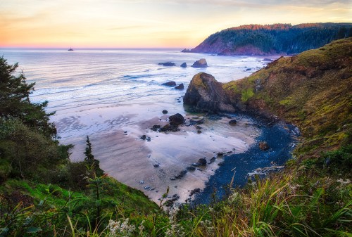 A scenic coastal view at sunset, featuring rocky cliffs, sandy beach, and gentle waves under a colorful sky.