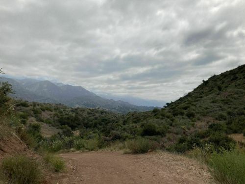 A dirt path winds through a green valley, surrounded by mountains under a cloudy sky.