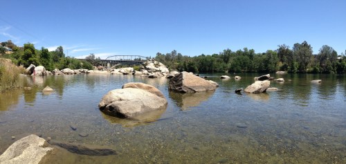 A serene river scene with large rocks, clear water, and a bridge in the background under a blue sky.