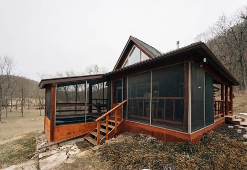 A rustic cabin with large screened porches, surrounded by trees and open land, on a cloudy day.