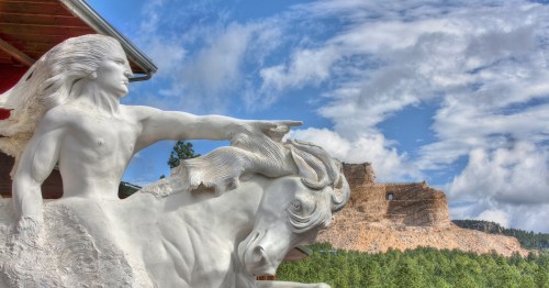 A white statue of a man on a horse points towards a distant rocky cliff under a blue sky with clouds.