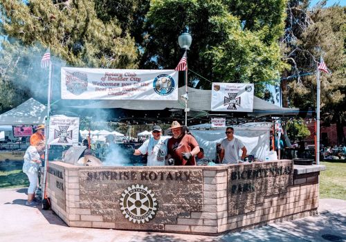 Three smiling men at a booth with banners for the Rotary Club's Beer Barrel Challenge at a park event.