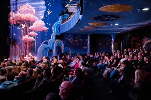 A vibrant theater audience seated in front of a colorful mural with clouds and whimsical designs on the walls.