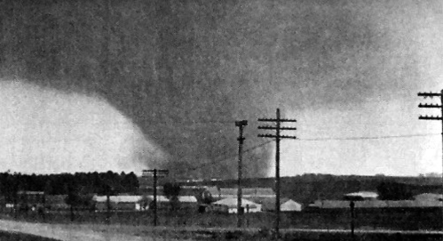 A dark tornado looms over a rural landscape with power lines and buildings in the foreground.