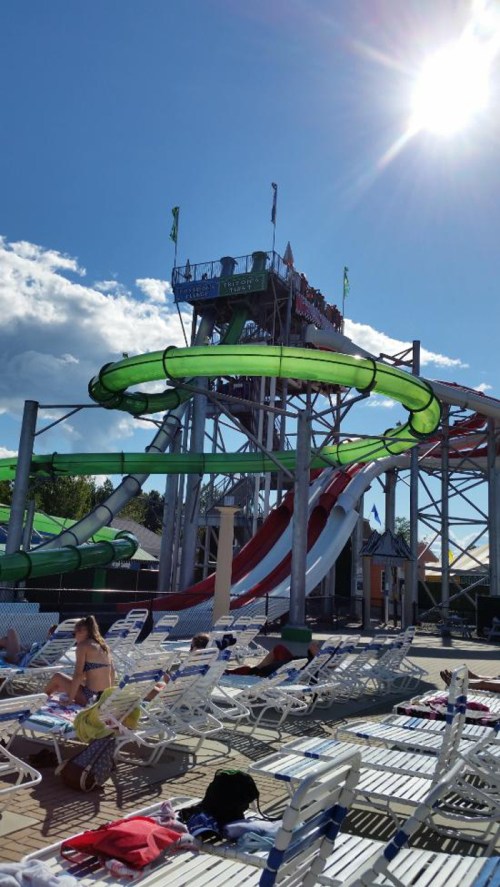 A water park featuring a tall slide structure with green and red slides, surrounded by sunbathers on lounge chairs.