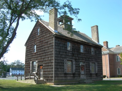 Historic wooden house with a shingled exterior, chimney, and a small cupola, set in a grassy area.