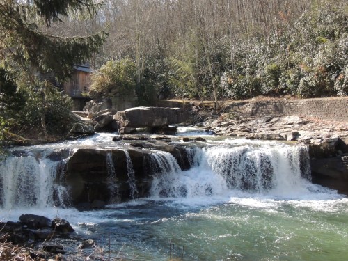 A serene waterfall cascades over rocks, surrounded by trees and a rustic building in the background.