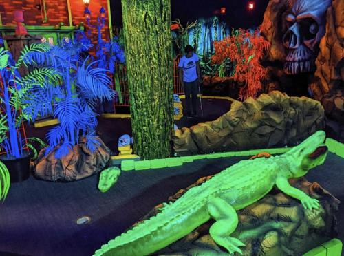 A vibrant mini-golf scene with neon plants, a large green alligator statue, and a child playing in the background.
