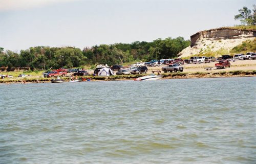 A view of a lake with boats and a beach area, surrounded by trees and parked vehicles in the background.