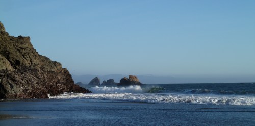 A serene beach scene with rocky cliffs, gentle waves, and distant rock formations under a clear blue sky.
