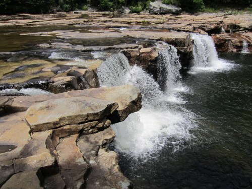 A serene waterfall cascading over rocky ledges into a clear pool, surrounded by lush greenery.