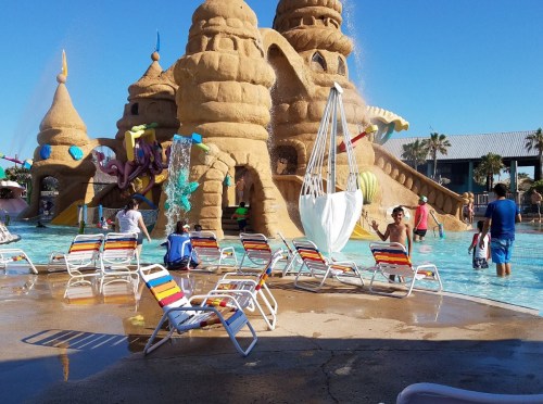 A water park scene with a sandy castle structure, colorful lounge chairs, and people enjoying the water on a sunny day.