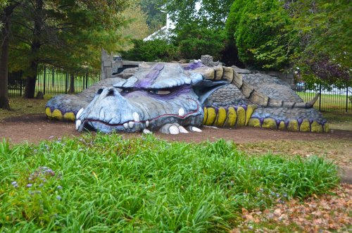 A large, colorful dragon sculpture lies on the ground, surrounded by greenery and trees in a park setting.