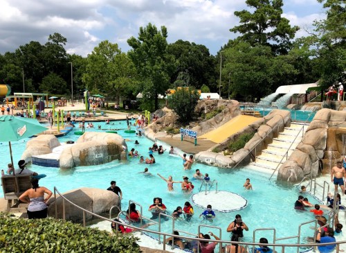 A busy water park with slides, pools, and families enjoying the sun and water activities. Lush greenery surrounds the area.