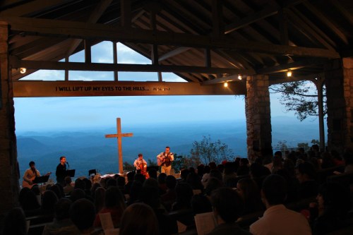 A worship service in a mountain chapel with a cross, overlooking a scenic view and a large audience.