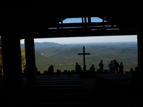 Silhouette of people inside a chapel, overlooking a mountainous landscape with a cross in the foreground.