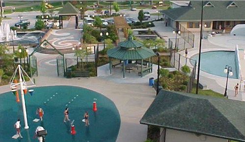 Aerial view of a community pool area with water features, a gazebo, and landscaped gardens. People are enjoying the space.