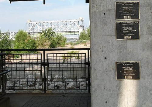 Plaques on a concrete wall near a river, with a bridge visible in the background and greenery surrounding the area.