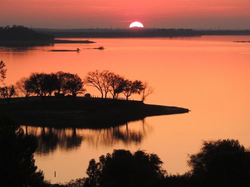 A serene sunset over a lake, with silhouettes of trees and a warm orange sky reflecting on the water.