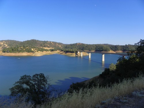A serene landscape featuring a bridge over a calm lake, surrounded by hills and trees under a clear blue sky.