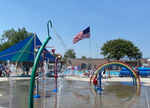 A splash pad with colorful water features, children playing, and an American flag waving in the background on a sunny day.