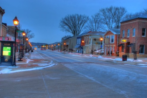 Quiet street scene at dusk, lined with historic buildings and lampposts, lightly dusted with snow.