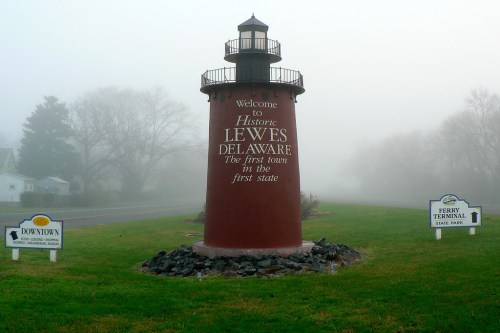 A foggy scene featuring a lighthouse sign welcoming visitors to Lewes, Delaware, with nearby directional signs.