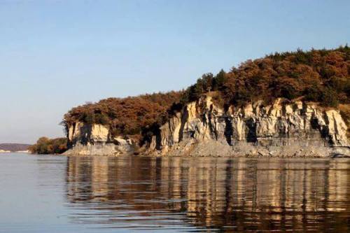 A serene landscape featuring a rocky cliff with autumn foliage, reflected in calm water under a clear sky.