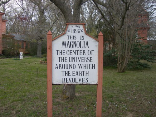 Sign reading "This is Magnolia, the center of the universe around which the Earth revolves," set in a grassy area.