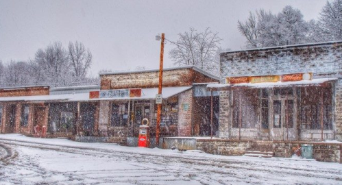 A snowy scene of an old, rustic storefront with a vintage gas pump, surrounded by trees and a winter landscape.