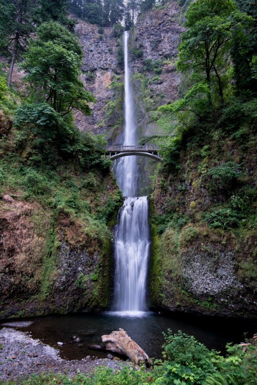 A stunning waterfall cascades down rocky cliffs, with a bridge spanning above and lush greenery surrounding the scene.