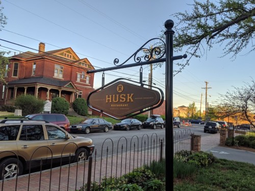 Sign for Husk Restaurant in front of a historic building, with parked cars and trees in the background.