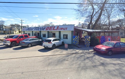 Colorful taco restaurant with outdoor seating, surrounded by parked cars on a sunny day.