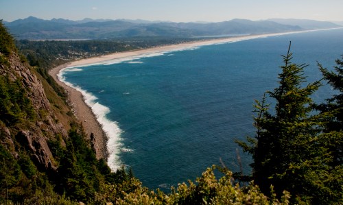 A scenic coastal view with waves crashing on a sandy beach, framed by lush greenery and distant mountains.